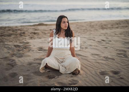 Corpo pieno di calma giovane etnia barefoted turista femminile con lunghi capelli ondulati scuri in abbigliamento casual riposante su spiaggia sabbiosa con gambe incrociate e guardare Foto Stock