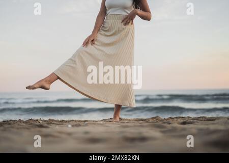 Viaggiatore femminile corto irriconoscibile con lunghi capelli scuri in estate casual indossare calci di sabbia e sorridente mentre si trova in piedi sulla spiaggia vicino oceano ondulato a s Foto Stock