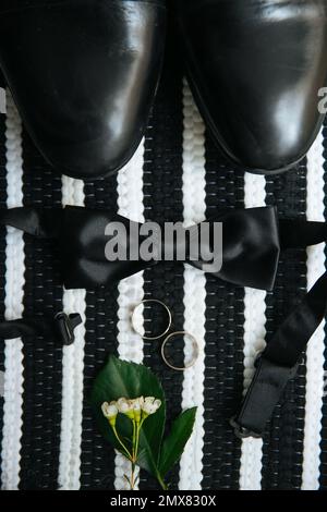 Vista dall'alto della cravatta nera e delle scarpe in pelle poste su sfondo bianco e nero vicino ad anelli di nozze e fiori delicati Foto Stock