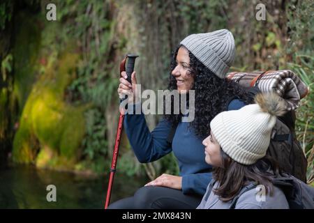 Madre etnica e figlia positiva in abiti caldi seduti sulla riva del lago con bastone da trekking mentre ammirano la natura durante il viaggio di trekking insieme Foto Stock