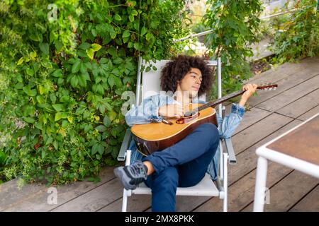 Bel giovane afro-italiano uomo di corsa mista in abbigliamento casual suonare la chitarra in giardino. Un ragazzo multirazziale suona uno strumento musicale riposa e. Foto Stock