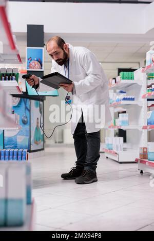 Farmacista che guarda le informazioni delle pillole mediche sul computer tablet mentre scansiona il codice a barre delle droghe usando lo scanner della farmacia. Il dipendente della farmacia è responsabile dell'organizzazione e dell'etichettatura dei pacchetti Foto Stock