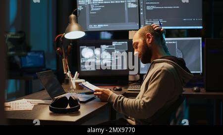 Tecnico IT maschile che analizza i dati web su appunti e schermate, confrontando le informazioni sui file. Giovane uomo che usa il linguaggio di programmazione e codifica la nuova interfaccia utente di notte. Foto Stock