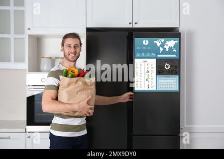 Giovane uomo con sacchetto di generi alimentari vicino smart frigorifero in cucina Foto Stock