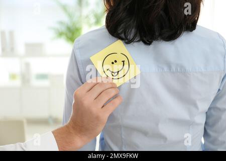 Uomo che mette divertente adesivo sorridente sul retro del collega in ufficio, primo piano. Aprile Fool's Day Foto Stock