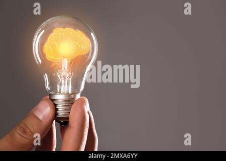 Uomo che tiene la lampadina con il cervello splendente all'interno contro lo sfondo grigio, spazio per il testo. Generazione di idee Foto Stock