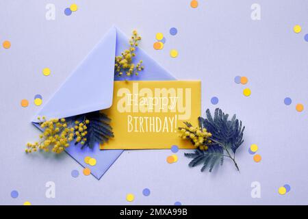 Testo Happy Birthday carta bianca viola chiaro, carta bianca gialla, fiori mimosa freschi in busta viola chiaro su sfondo lilla con carta Foto Stock