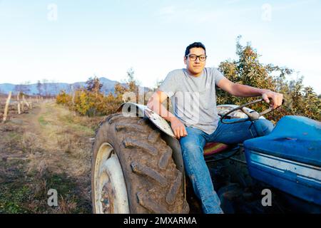 Vista frontale di un contadino latino che guida un vecchio trattore, nel mezzo della sua campagna al tramonto Foto Stock