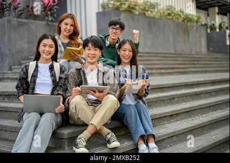 Gruppo di giovani studenti asiatici felici in abiti casual seduti sulle scale di fronte all'edificio del campus, sorridendo e guardando la macchina fotografica. te Foto Stock