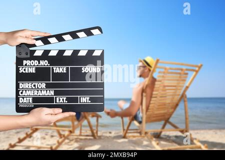 Assistente che tiene il clapperboard e le persone che si rilassano in sedie a sdraio sulla spiaggia, primo piano. Produzione cinematografica Foto Stock