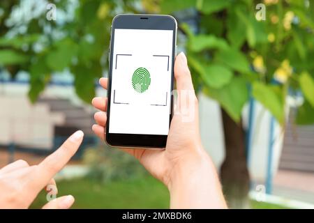 Woman holding smartphone with fingerprint sensor outdoors, closeup. Digital identity Foto Stock
