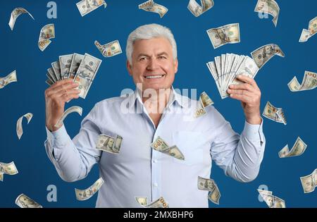 Uomo anziano con dollari americani sotto la pioggia di soldi su sfondo blu Foto Stock