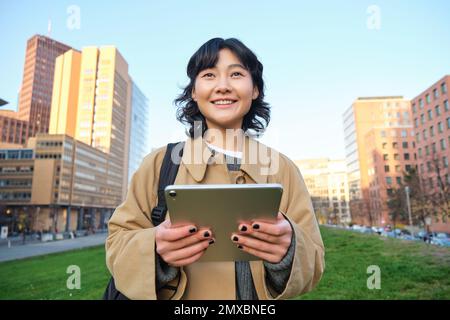 Felice ragazza asiatica si trova sulla strada, studenti universitari cammina con tablet digitale in mani e sorrisi, si trova nel centro della città Foto Stock