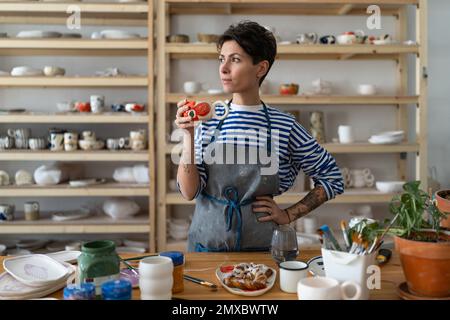 Donna ceramista creativa in grembiule sporco godendo il suo caffè mattutino dalla tazza di argilla e guardando via Foto Stock