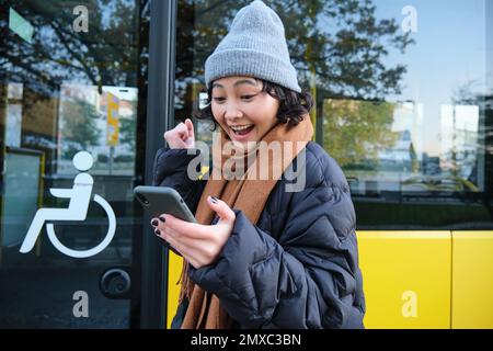 Persone moderne e stile di vita. Felice ragazza asiatica urla da gioia, festeggia, si trova vicino ai trasporti pubblici degli autobus e sembra stupito Foto Stock