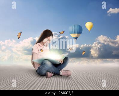Carino bambina lettura libro magico. Vista pittoresca del cielo blu e balloni d'aria sullo sfondo Foto Stock