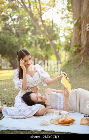 Ritratto, due belle e affascinanti donne asiatiche godono di picnic insieme nel giardino nei fine settimana, una donna che si trova sui giri del suo amico durante la lettura Foto Stock