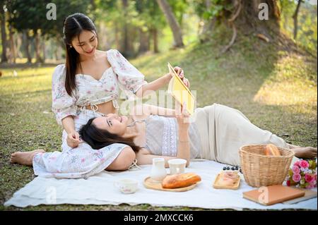 Due belle donne asiatiche amano fare picnic insieme nel giardino nei fine settimana, una donna che si trova sui giri dell'amico mentre legge un libro. Foto Stock