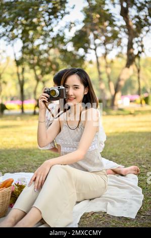 Ritratto di bella bella donna asiatica in abiti casual si siede sul vestito da picnic, tenendo una fotocamera retrò, sorridendo e guardando nella fotocamera. Foto Stock