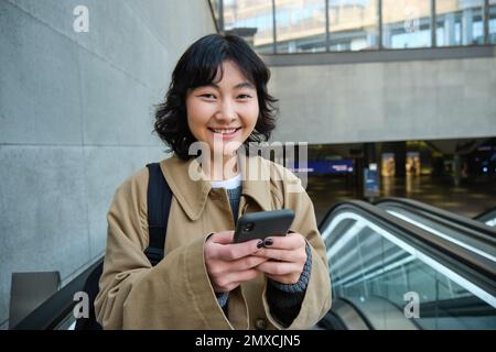 Giovane donna bruna si sposta, va da qualche parte in città, si trova su scala mobile e utilizza il telefono cellulare, tiene lo smartphone e sorride. Foto Stock