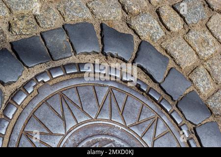 Vecchio portello della fogna del metallo nel mezzo della strada, disposto da una pietra di pavimentazione liscia. Foto Stock