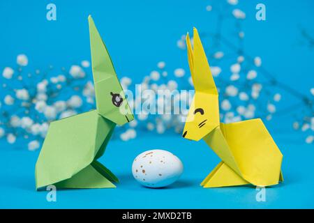 Due coniglietti di origami di carta e un uovo di Pasqua su sfondo blu. Artigianato con le proprie mani per Pasqua Foto Stock