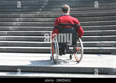 utente su sedia a rotelle in piedi di fronte a una scala esterna non priva di barriere architettoniche, vista posteriore Foto Stock