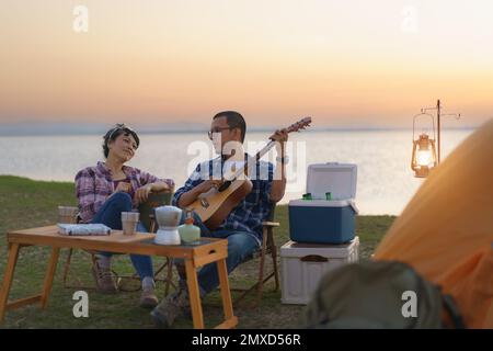 Coppia asiatica che canta e suona la chitarra nel loro campeggio con lago sullo sfondo durante il tramonto. Foto Stock
