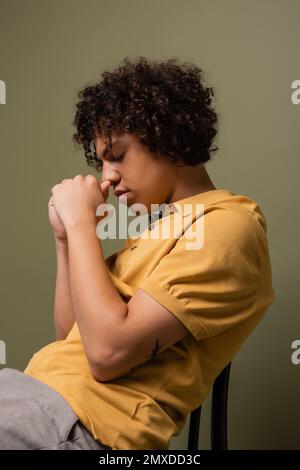 vista laterale dell'uomo afroamericano in polo giallo seduto su sedia con gli occhi chiusi e le mani vicino al viso su sfondo grigio verde, immagine stock Foto Stock