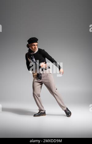 intera lunghezza dell'uomo afroamericano in berretto elegante e maglione nero con sciarpa a scacchi che guarda via mentre posa su sfondo grigio, immagine di scorta Foto Stock