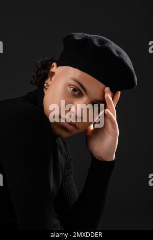 ritratto di elegante afro-americano ragazzo in beretta e dolcevita tenendo la mano vicino al viso e guardando la fotocamera isolato su nero, immagine stock Foto Stock