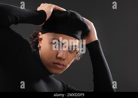 giovane e trendy afroamericano uomo in nero dolcevita regolazione beretta e guardando la macchina fotografica isolato su grigio scuro, immagine stock Foto Stock