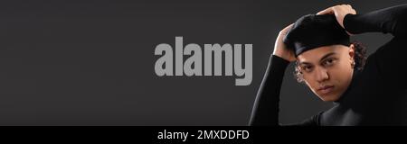 giovane uomo afroamericano in maglione nero regolazione berretto elegante mentre si guarda la fotocamera isolato su grigio scuro, banner, immagine stock Foto Stock