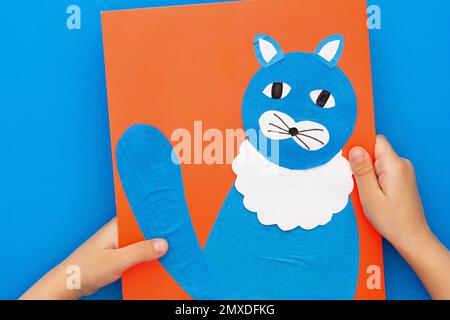 Vista dall'alto delle mani dei bambini che tengono gatto di carta artigianale su foglio rosso su sfondo blu Foto Stock