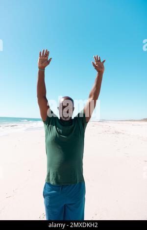 Uomo afroamericano calvo anziano con le braccia sollevate meditando sulla spiaggia sabbiosa sotto il cielo chiaro Foto Stock