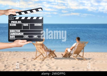 Assistente che tiene il clapperboard e le persone che si rilassano in sedie a sdraio sulla spiaggia, primo piano. Produzione cinematografica Foto Stock