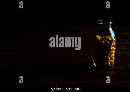 Capsule gialle profonde in bottiglia di vetro trasparente su tavolo di legno con illuminazione a basso contenuto di tasti. Foto Stock