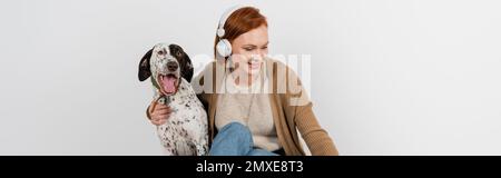 Donna sorridente in cuffie wireless che abbraccia cane dalmata a casa, banner, immagine stock Foto Stock