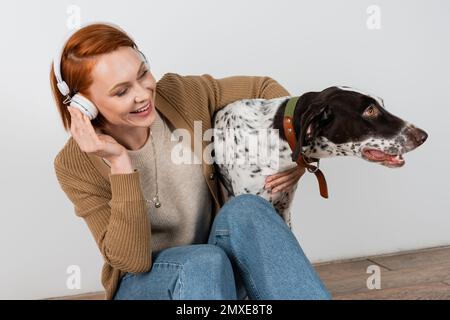 Allegra donna rossa che ascolta musica in cuffie e abbraccia cane dalmata a casa, immagine stock Foto Stock