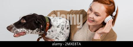 Donna positiva in cuffie abbracciando cane dalmata isolato sul grigio, banner, immagine stock Foto Stock