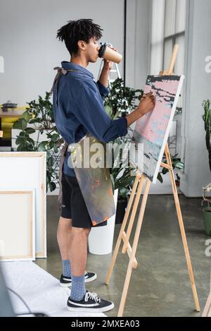 Vista laterale del giovane artista afro-americano bere caffè e pittura su tela in studio, immagine stock Foto Stock
