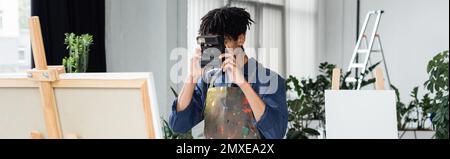 Artista afro-americano che scatta foto su una macchina fotografica vintage vicino a canvas su cavalletto in studio, banner, immagine stock Foto Stock