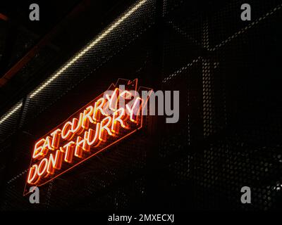 Iscrizione neon mangiare curry non fretta, attirando i visitatori di cucina indiana, colore rosso arancio, sfarfallio si illumina nella parete scura del ristorante. Foto Stock