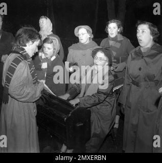 1955, storico, canto di carol, un gruppo di donne, alcuni più giovani, forse studenti universitari, alcuni più anziani, in cappotti e sciarpe, riuniti all'esterno intorno a un piccolo pianoforte cantando carole di Natale, due torce in mano, Cambridge, Inghilterra, Regno Unito, Foto Stock