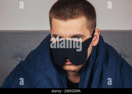 Giovane uomo imbarazzato in una coperta a casa indossa una maschera di sonno mentre si rilassa a casa, ritratto studio. Relax, stile di vita di buon umore Foto Stock