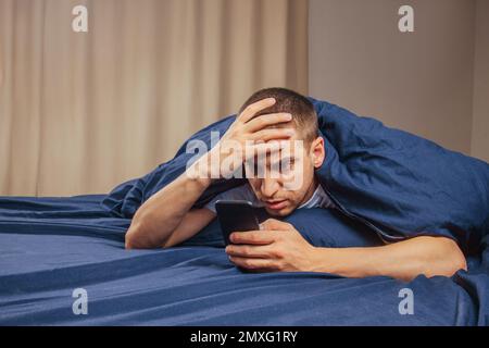 Cattive notizie, problemi, offerte e pubblicità. Scioccato ragazzo attraente sconvolto legge un messaggio sul telefono e preme la mano al suo volto su un letto bianco Foto Stock