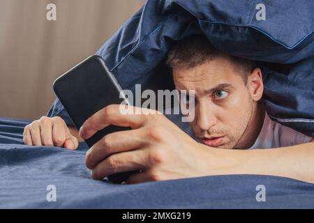 Cattive notizie, problemi, offerte e pubblicità. Scioccato ragazzo attraente sconvolto legge un messaggio sul telefono e preme la mano al suo volto su un letto bianco Foto Stock