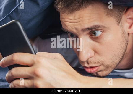 Cattive notizie, problemi, offerte e pubblicità. Scioccato ragazzo attraente sconvolto legge un messaggio sul telefono e preme la mano al suo volto su un letto bianco Foto Stock