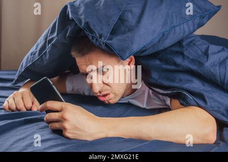 Cattive notizie, problemi, offerte e pubblicità. Scioccato ragazzo attraente sconvolto legge un messaggio sul telefono e preme la mano al suo volto su un letto bianco Foto Stock