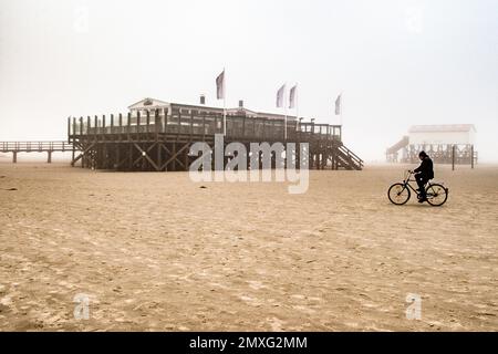Un ciclista attraversa la spiaggia sabbiosa nebbiosa di Sankt Peter-Ording, Germania, passando davanti a un ristorante su palafitte con diversi pali di bandiera sulla sua terrazza. Un secondo edificio su palafitte, più piccolo, è visibile nella foschia a destra. Foto Stock